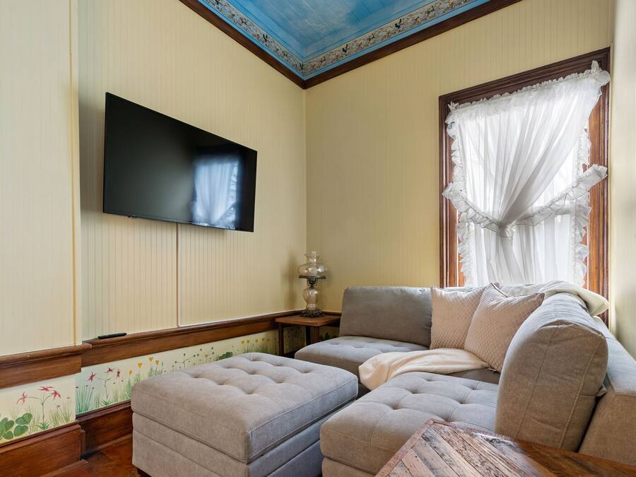 A living room with a sectional sofa, a flat-screen TV mounted on the wall, and a window with sheer curtains.