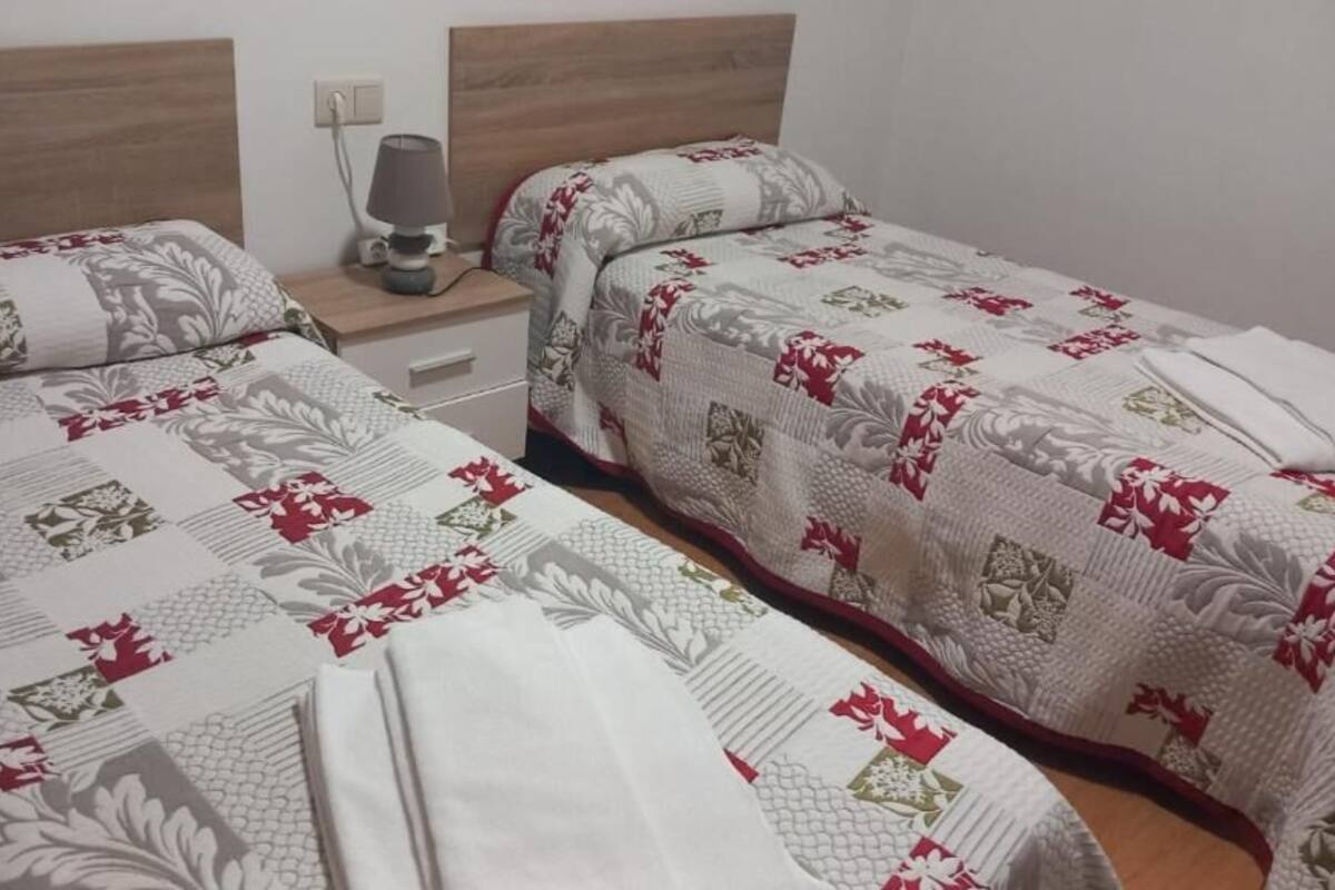 A double room with two beds, each with a floral-patterned bedspread. There is a bedside table with a lamp and a power outlet on the wall.