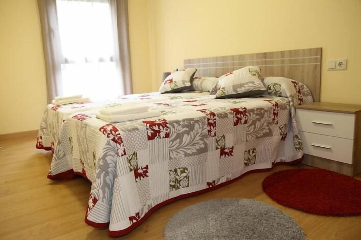 A neatly made bed with floral bedding, a bedside table, and a red rug on a wooden floor.