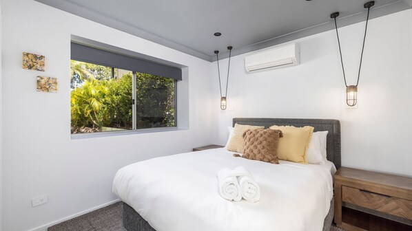 Une chambre moderne avec un grand lit, une fenêtre donnant sur un paysage verdoyant, un climatiseur fixé au mur et une table de chevet avec une lampe.