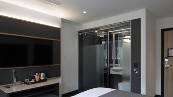 A modern hotel room with a large bed, a desk with a flat-screen TV, a glass shower enclosure, and a wooden door.