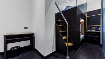 Quarto de hotel moderno com piso de madeira escura, uma escrivaninha com um cesto de lixo, uma escada e uma pequena área de estar com um banco.