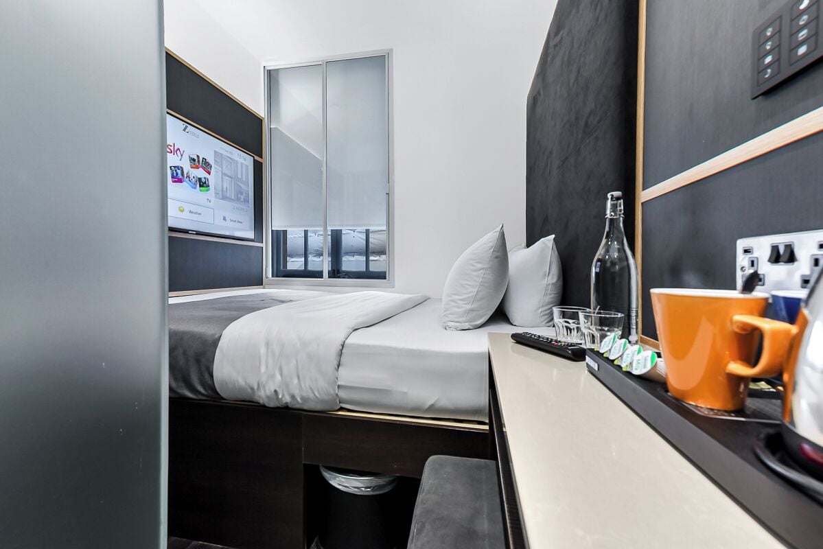 Un dormitorio pequeño con cama, una mesita con una botella, vasos y una taza, y un televisor mostrando una guía de canales.
