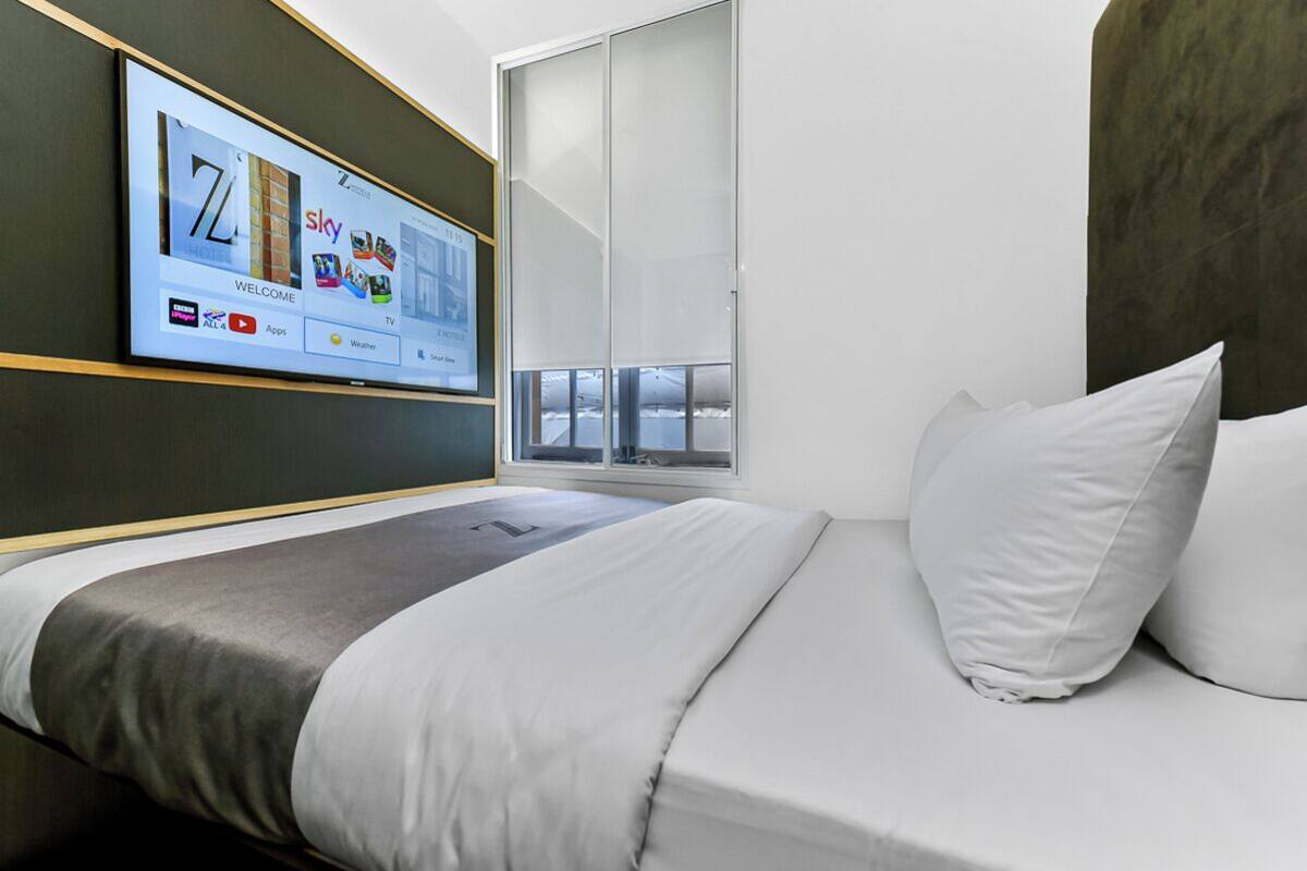 Een moderne hotelkamer met een grote flatscreen-tv waarop een hotelinterface wordt weergegeven, een bed met wit en grijs beddengoed, en een raam met uitzicht op de lucht.