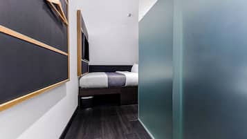 A narrow, modern room with a bed, a small bench, and a wall-mounted rack.