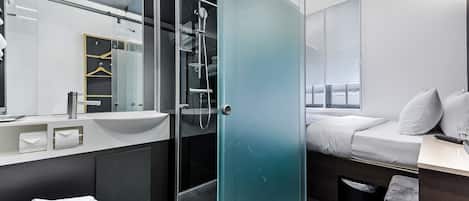 Una habitación de hotel moderna con cabina de ducha de vidrio, un espejo grande, una cama con ropa blanca y un pequeño escritorio con lámpara.