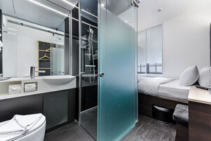 A modern hotel room with a glass shower enclosure, a large mirror, a bed with white bedding, and a small desk with a lamp.
