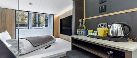 A modern hotel room with a bed, a desk with a kettle, and a window with a view of the outside.