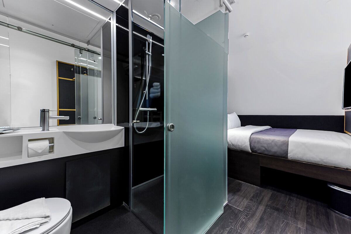 A modern hotel room with a glass shower enclosure, a bedside table, a bed, a sink, and a mirror.