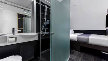 A modern hotel room with a glass shower enclosure, a bedside table, a bed, a sink, and a mirror.