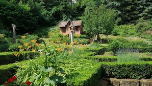 Un jardín con una pequeña casa de ladrillo rodeada de setos recortados y diversas plantas con flores.