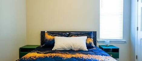 A bed with a blue quilt, a white pillow, and a green nightstand with a lamp.