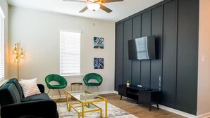 A modern living room with a dark wall panel, a flat-screen TV, a green chair, a black sofa, and a glass coffee table. - Stylish and NEW! Duke~Art Deco (Durham)