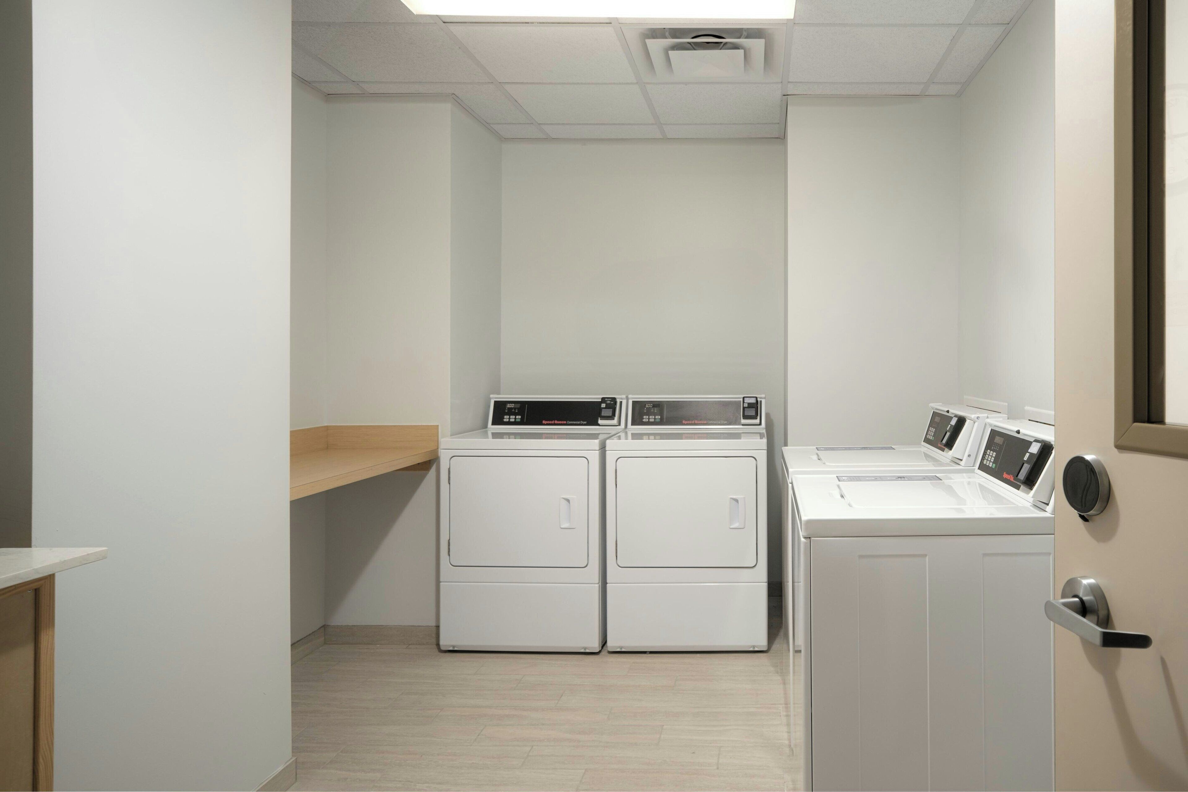 Une buanderie équipée de deux machines à laver et de deux sèche-linge à chargement frontal, d’une étagère en bois et d’une porte avec une poignée.