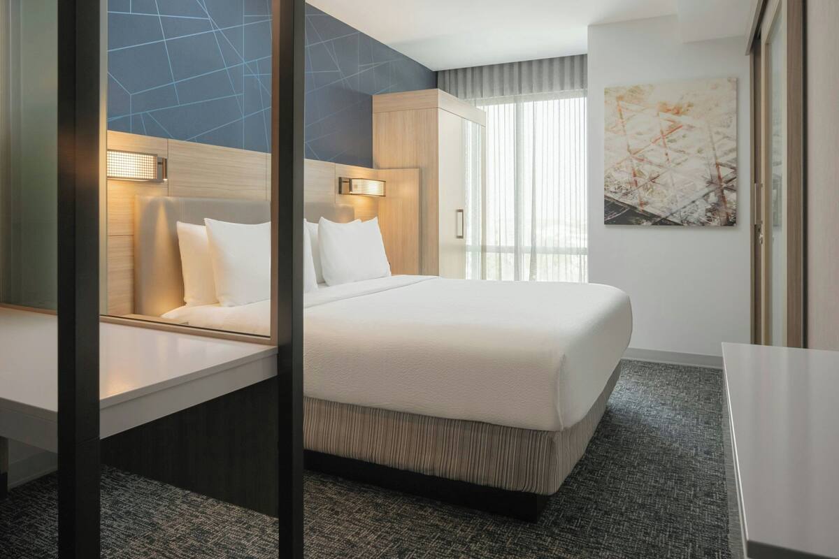 A modern hotel room with a large bed, a bedside table, a desk, and a large window with curtains.