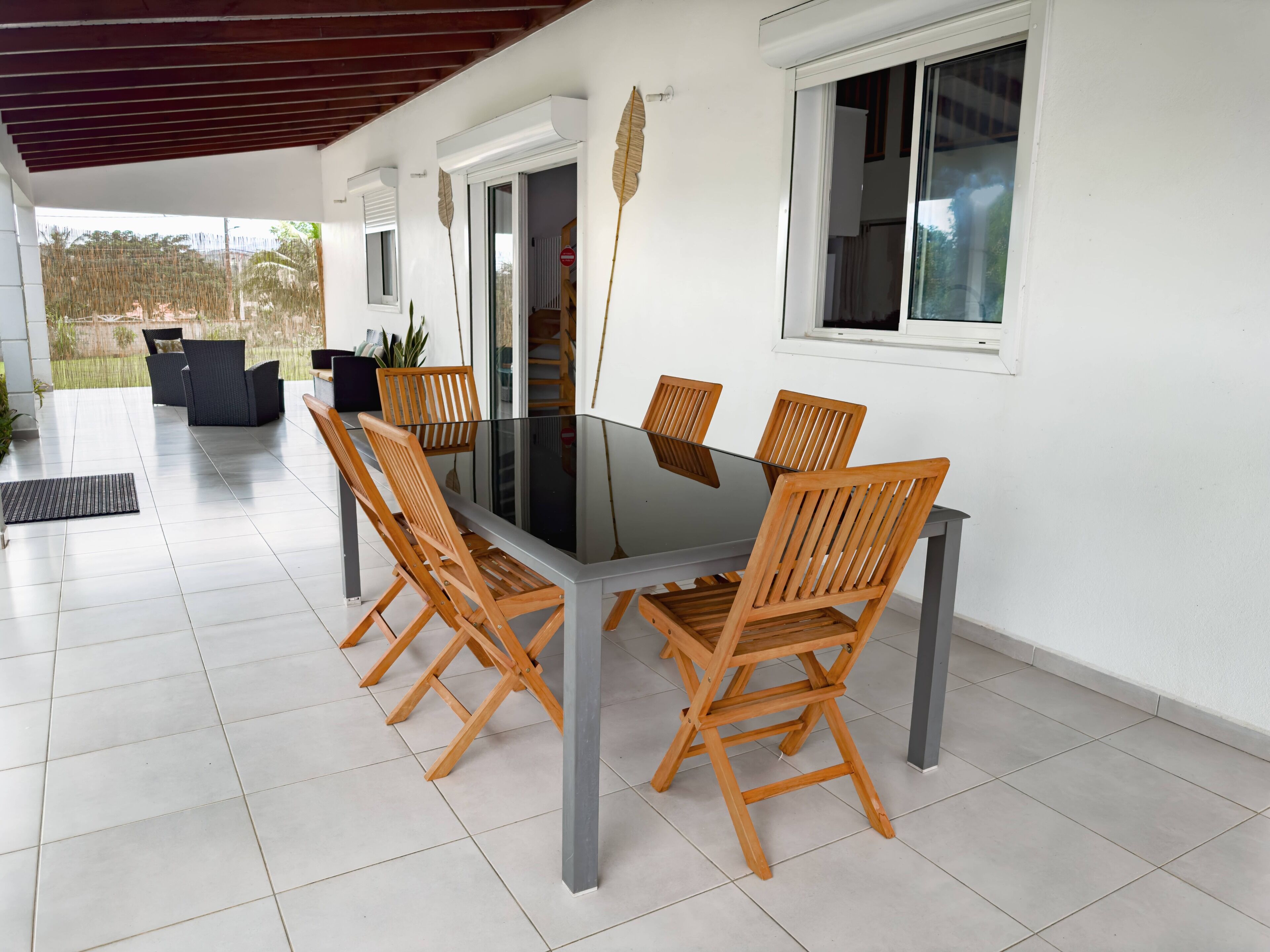 A modern outdoor dining area with a glass table and wooden chairs, surrounded by white walls and large windows.