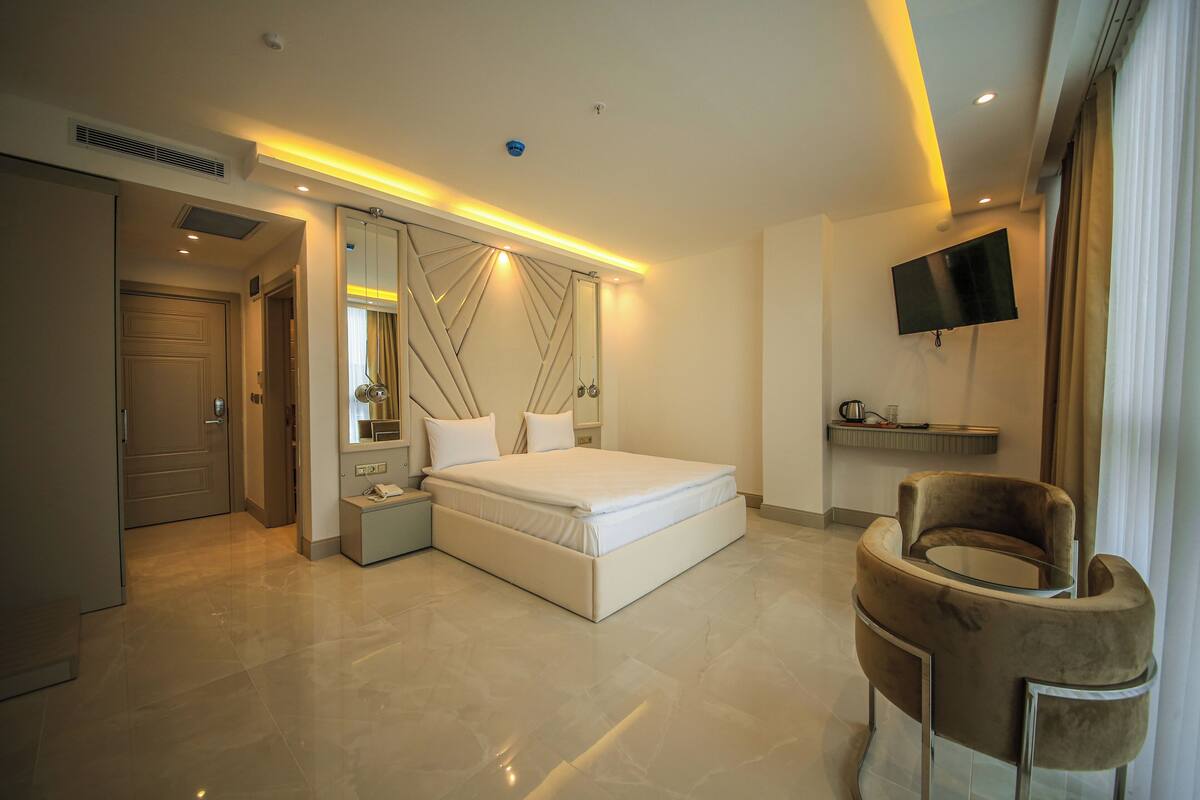 A modern hotel room with a large bed, a bedside table, a chair, a TV, and a bathroom door.