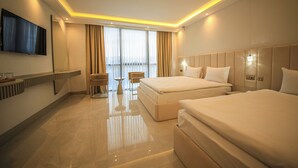 A modern hotel room with two beds, a flat-screen TV mounted on the wall, a desk with a chair, and large windows with curtains. - GÖRGÜLÜ HOTEL (Kesan)