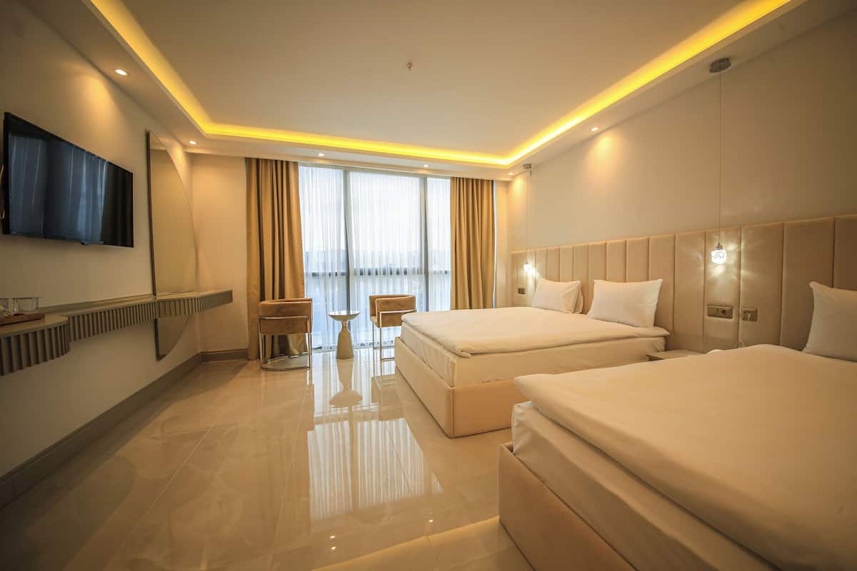 A modern hotel room with two beds, a flat-screen TV mounted on the wall, a desk with a chair, and large windows with curtains.