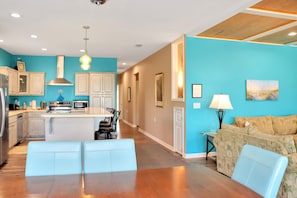 A modern kitchen with white cabinets, a central island, and a dining area with blue chairs. - Dolphin Daze Beach House (Port St. Joe)