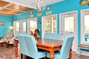 A dining area with a wooden table and light blue chairs, a chandelier, and a view of the outdoors through large windows. - Dolphin Daze Beach House (Port St. Joe)