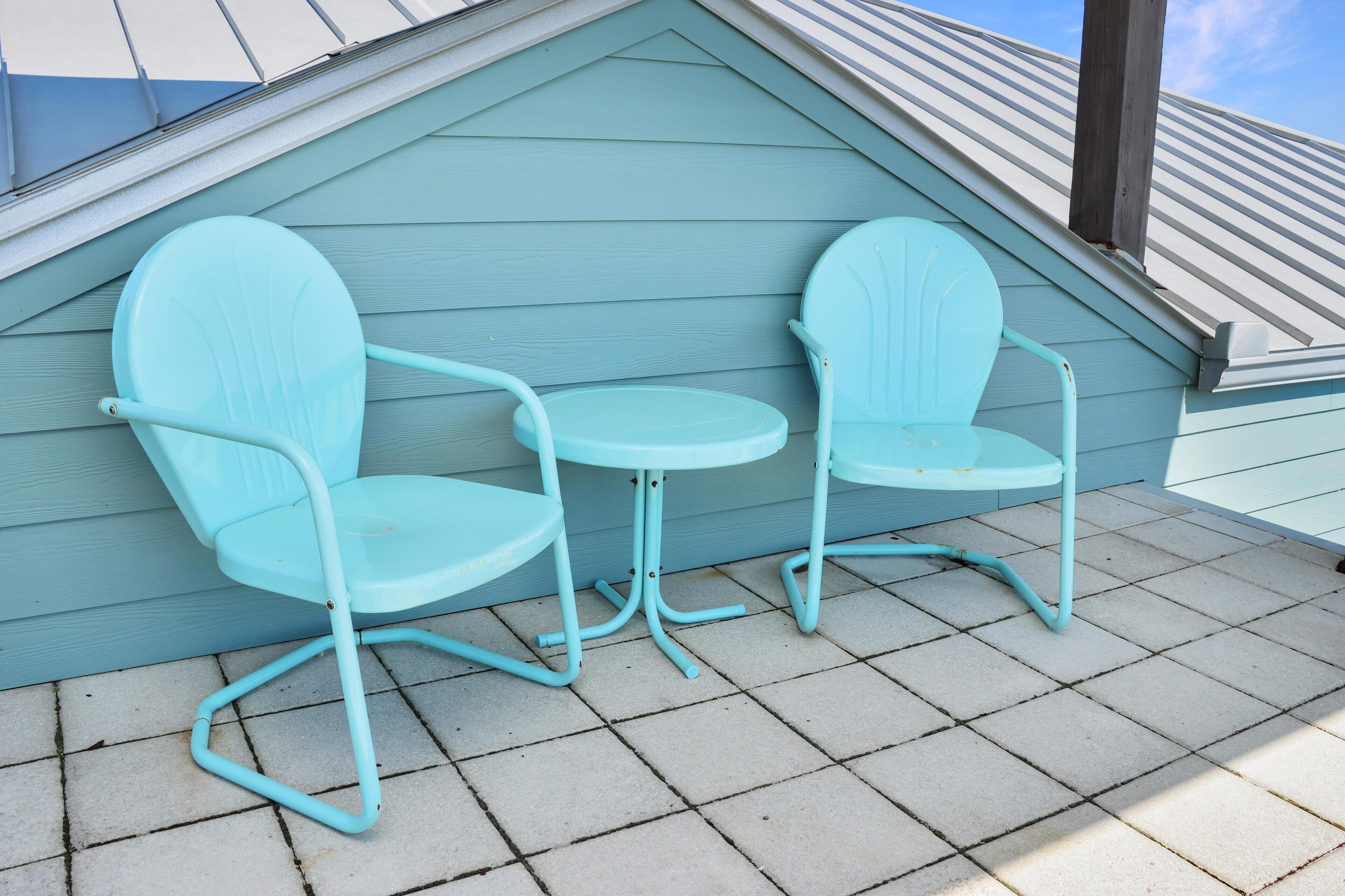 Un pequeño espacio al aire libre con dos sillas y una mesa frente a una pared azul claro, sobre un patio con baldosas.