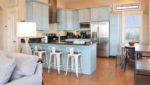 A modern kitchen with light blue cabinets, a central island with bar stools, stainless steel appliances, and a dining area with a wooden table and chairs. - The Blue Turtle (Port St. Joe)