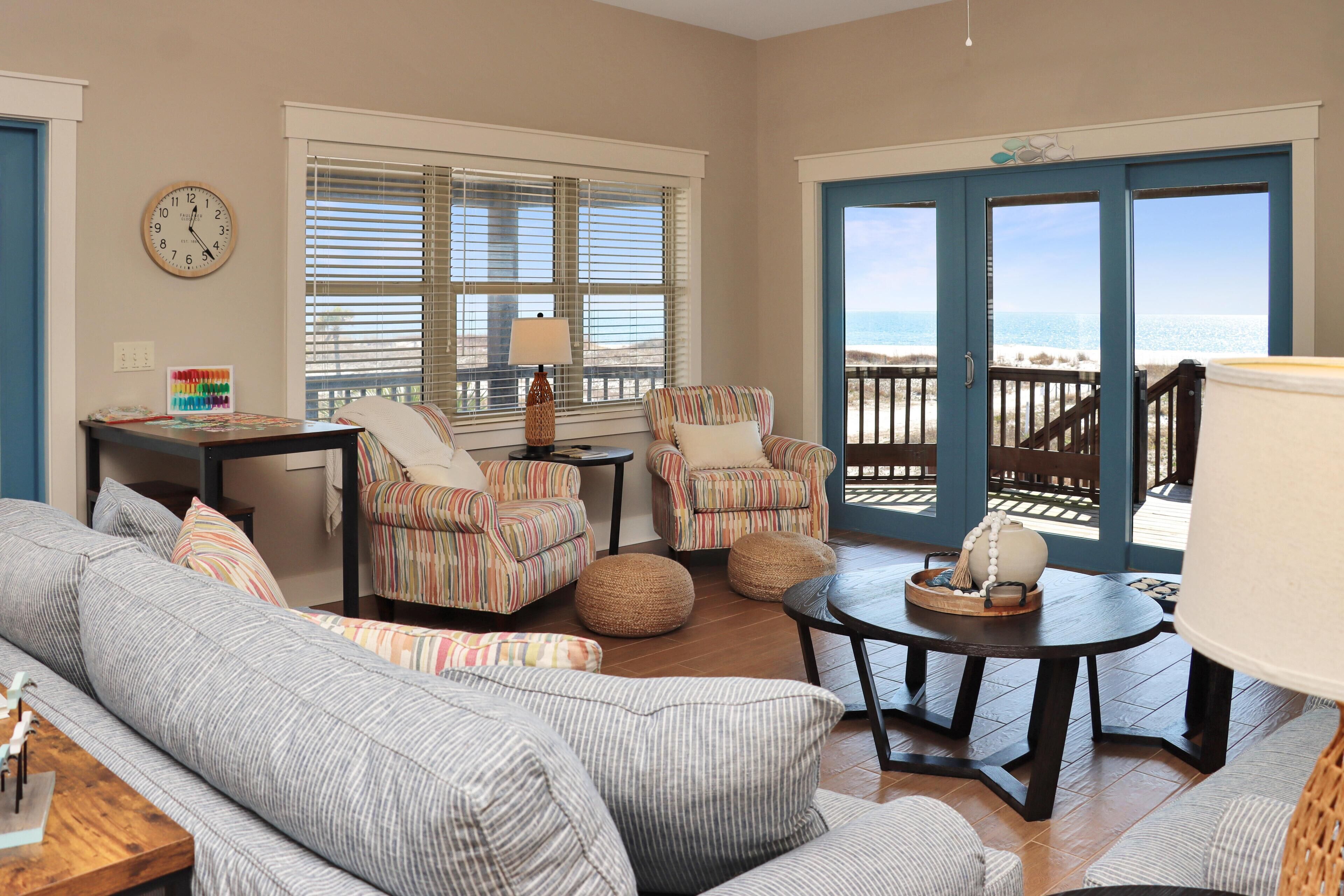 A living room with striped sofas, armchairs, and a coffee table. There is a clock on the wall and a view of the beach through large doors.