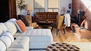 Un salon chaleureux avec un haut plafond en bois, une cheminée et une grande fenêtre équipée de stores.