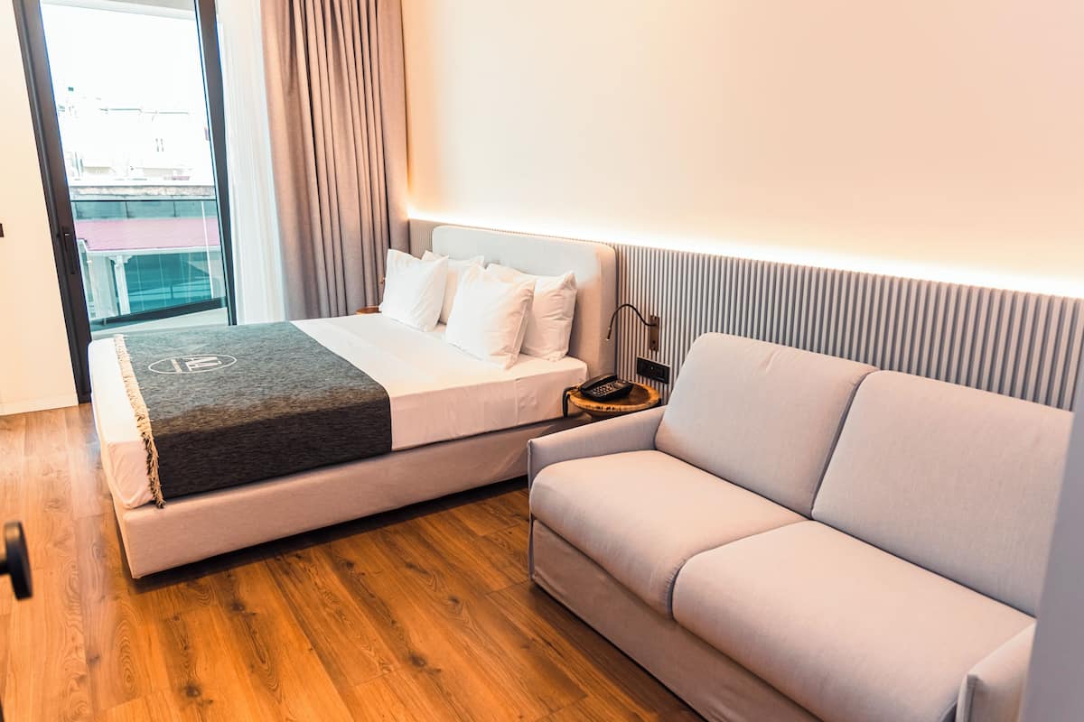 A modern hotel room with a bed, a sofa, a bedside table with a lamp, and a view through a window.