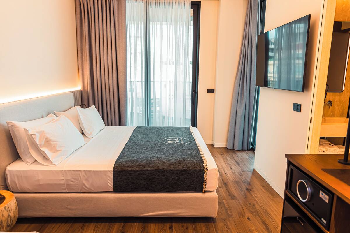 A neatly made bed with white linens and a dark throw blanket, a flat-screen TV mounted on the wall, and a wooden dresser with a circular handle.