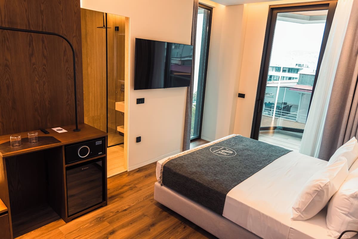 A hotel room with a bed, a TV mounted on the wall, a wooden wardrobe, and a small kitchenette.