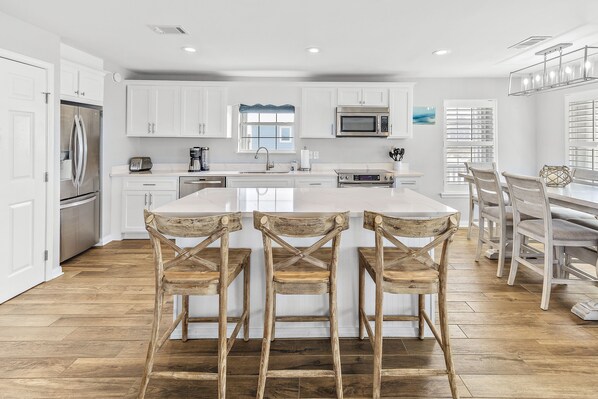 A modern kitchen with white cabinets, a stainless steel refrigerator, a microwave, and a large island with bar stools. - 50 Steps to the Beach with Ocean View ~ Beach Gear (Gulf Shores)