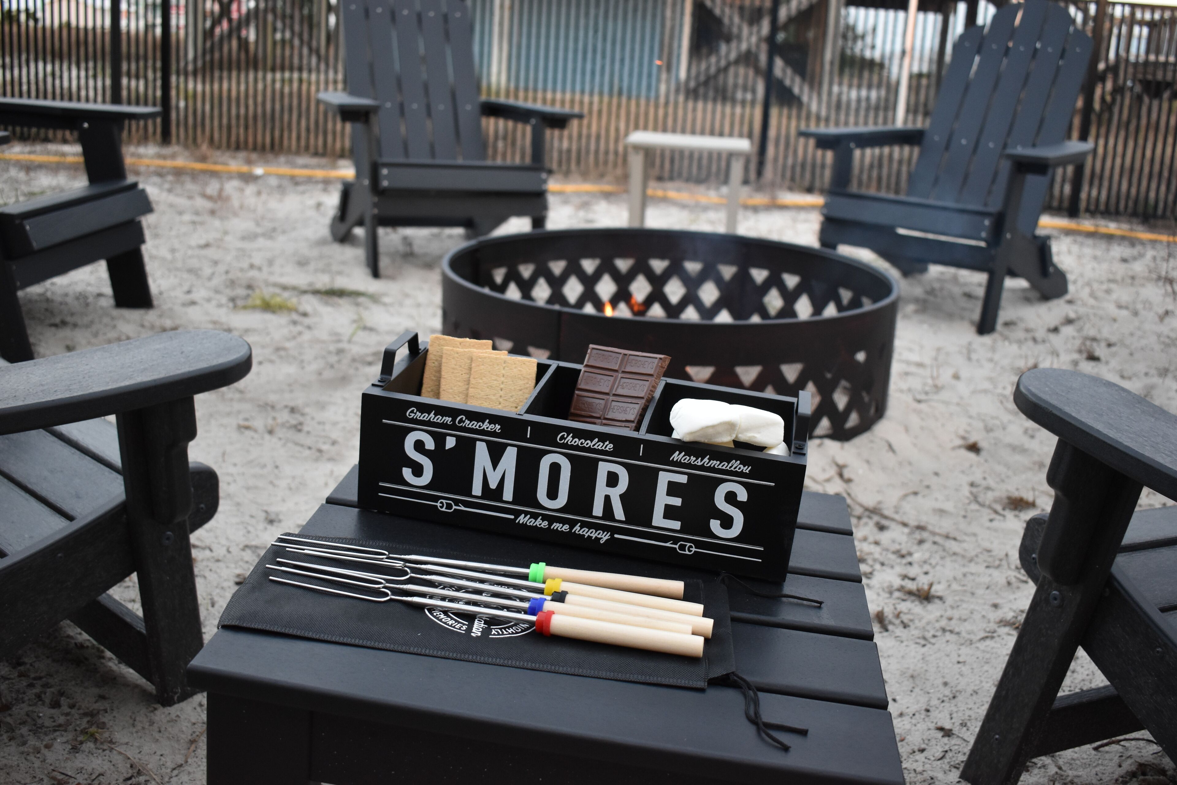 Caixa preta com o rótulo 'S'MORES', marshmallows, chocolate e palitos para s'mores sobre uma mesa em área externa arenosa, com cadeiras pretas e um braseiro ao fundo.
