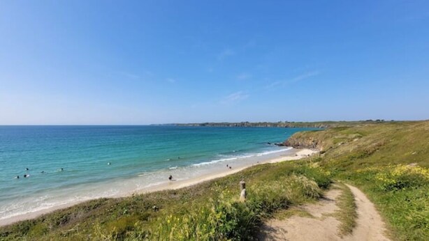 En kyststrand med en klar sti ned til vandkanten, omgivet af grønne områder og en fjerntliggende kystlinje.