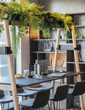 Un espace de salle à manger moderne, avec une longue table, des chaises et un arrangement décoratif de plantes au-dessus.