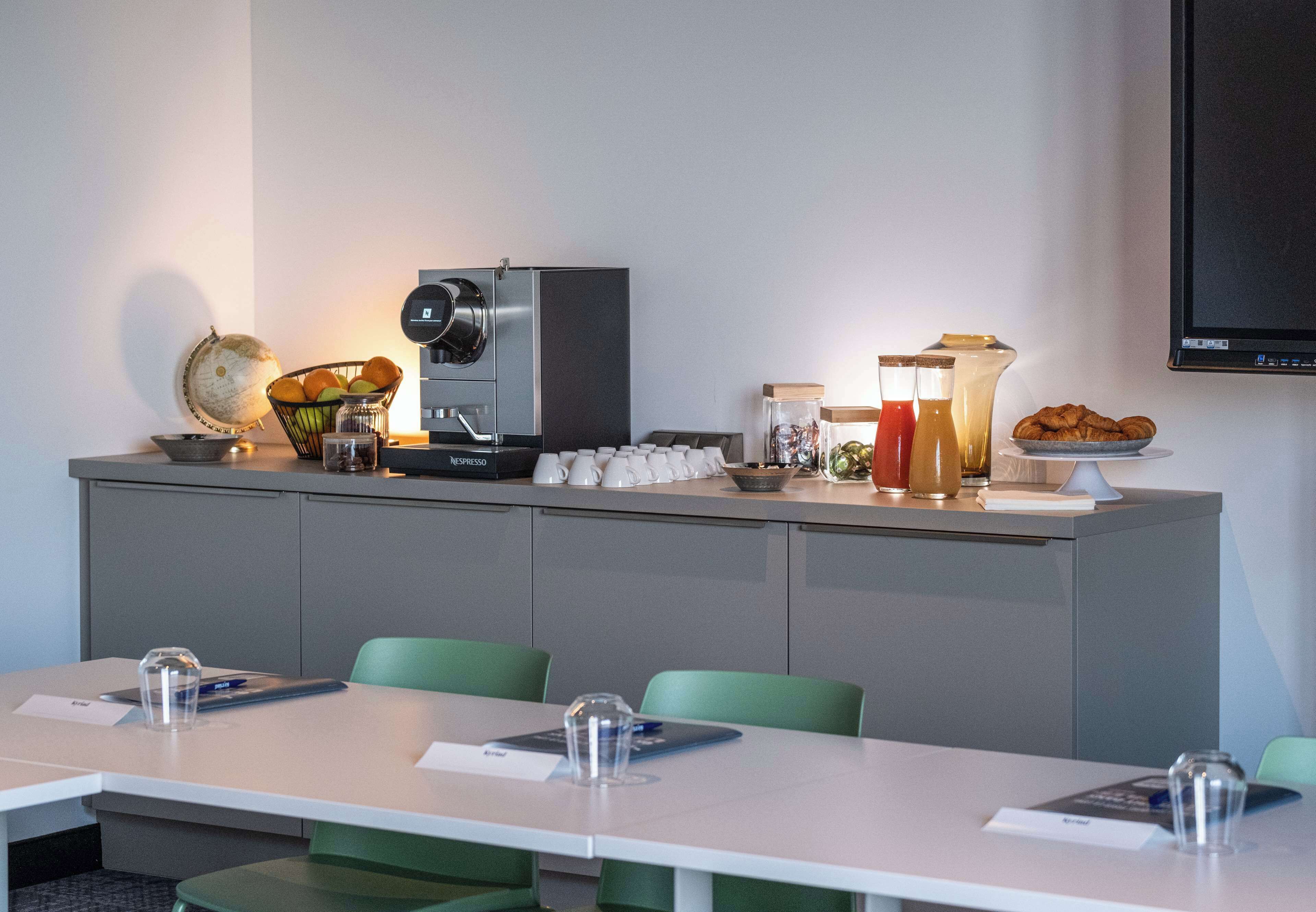 A modern hotel room with a kitchenette, a dining area with a long table, and a flat-screen TV mounted on the wall.