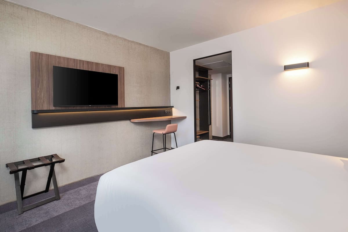 Een moderne hotelkamer met een groot bed, een flatscreen televisie bevestigd aan een houten paneel, een klein bureau met stoel, en een inloopkast.
