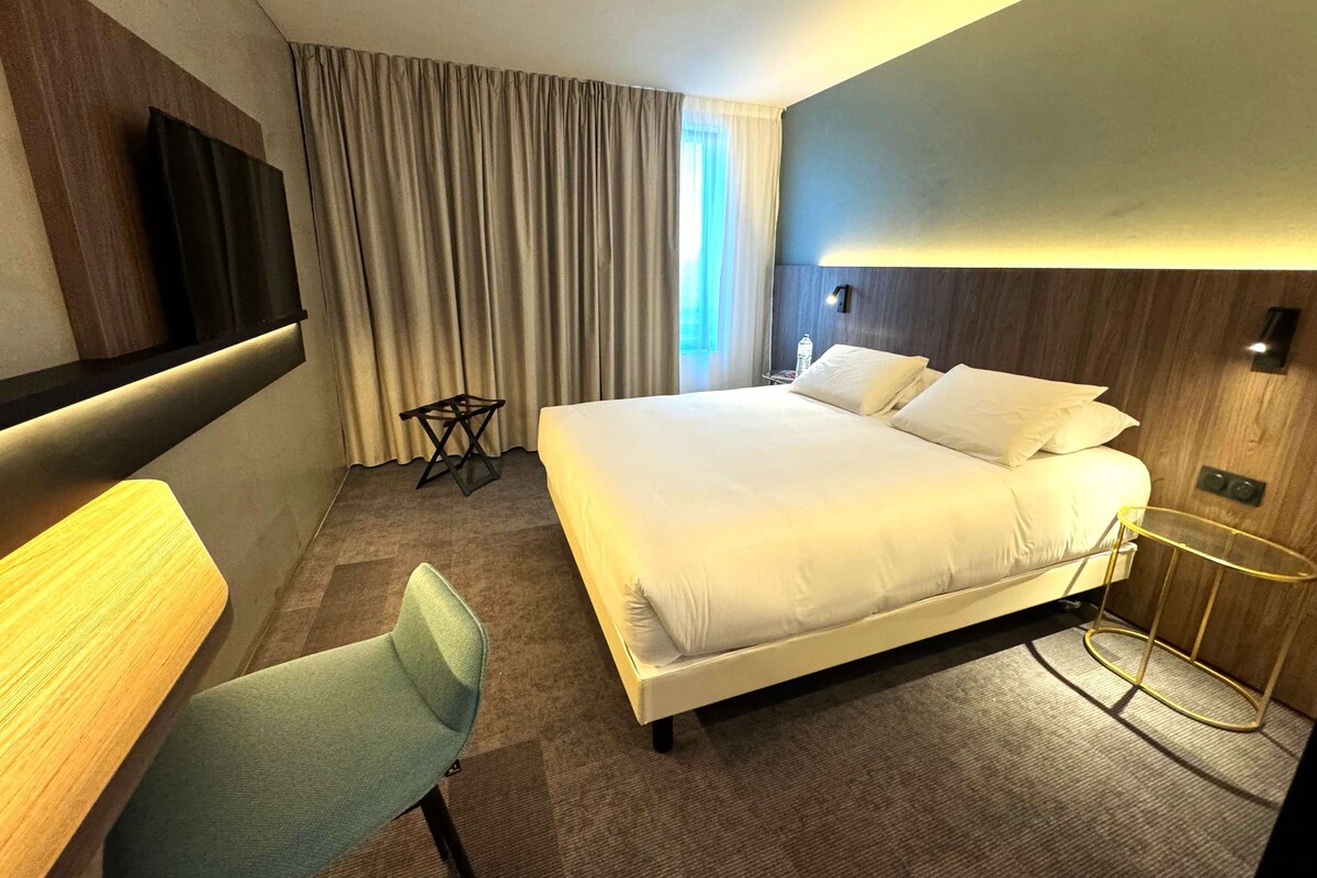 A hotel room with a large bed, a desk, a chair, a TV, and a window with curtains.