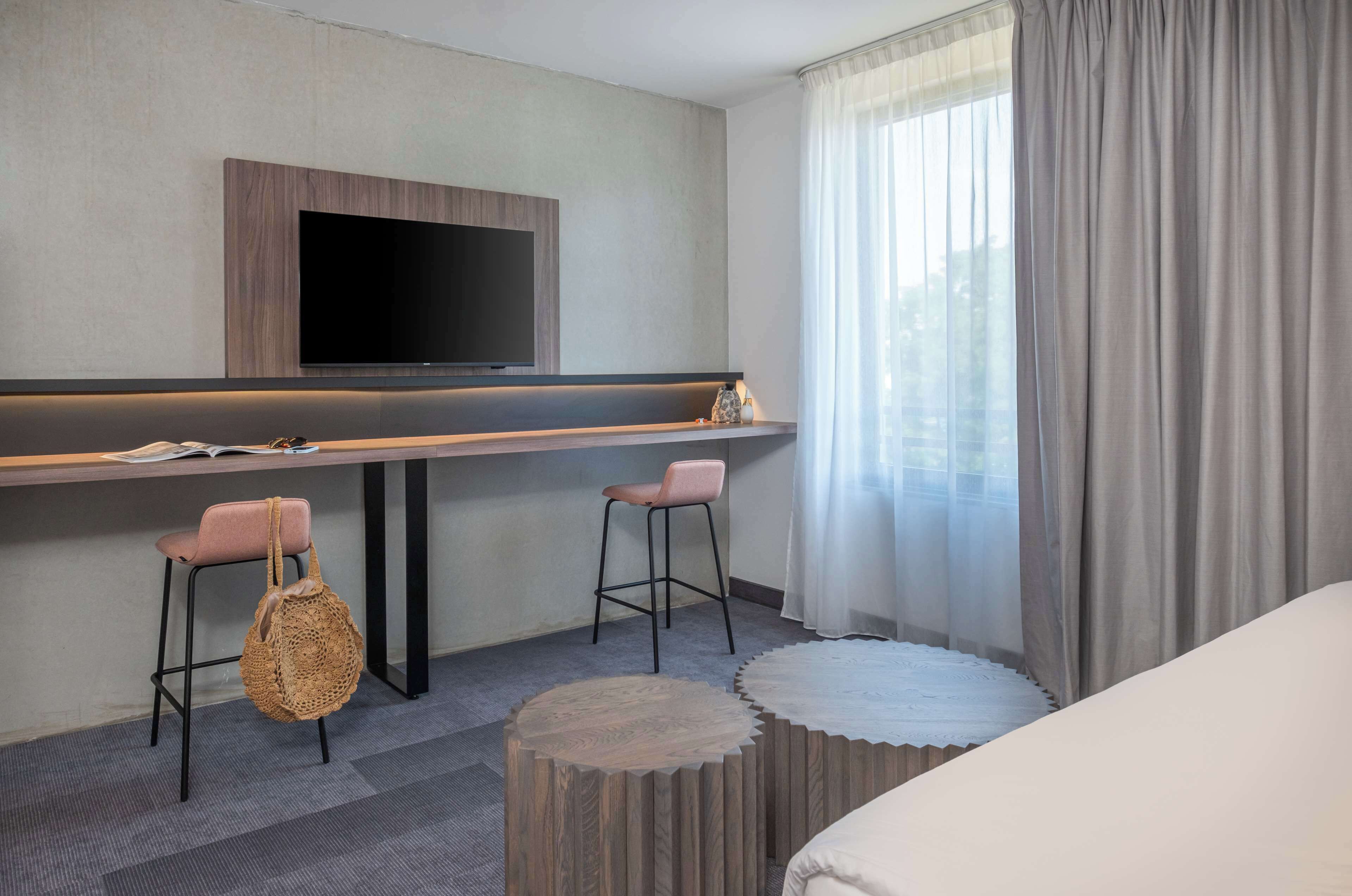 A modern hotel room with a flat-screen TV mounted on the wall, two bar stools, a round wooden side table, and a large window with curtains.