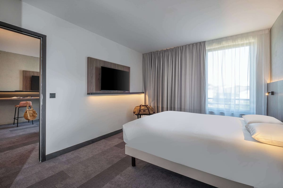 Een hotelkamer met een groot bed, een flatscreen televisie aan de muur, een raam met gordijnen en een klein tafeltje met een stoel en een handtas.