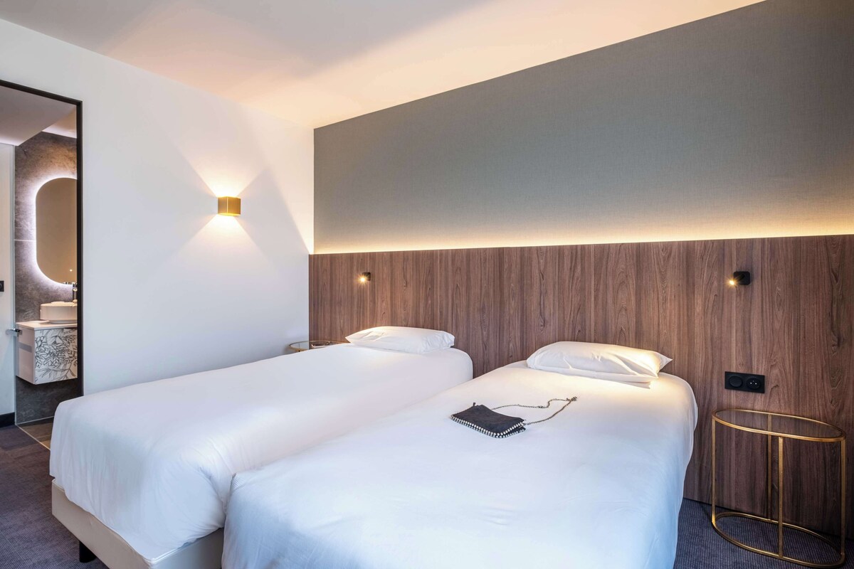 Une chambre d’hôtel avec deux lits, une tête de lit en bois, une table de chevet ronde dorée et un luminaire fixé au mur.