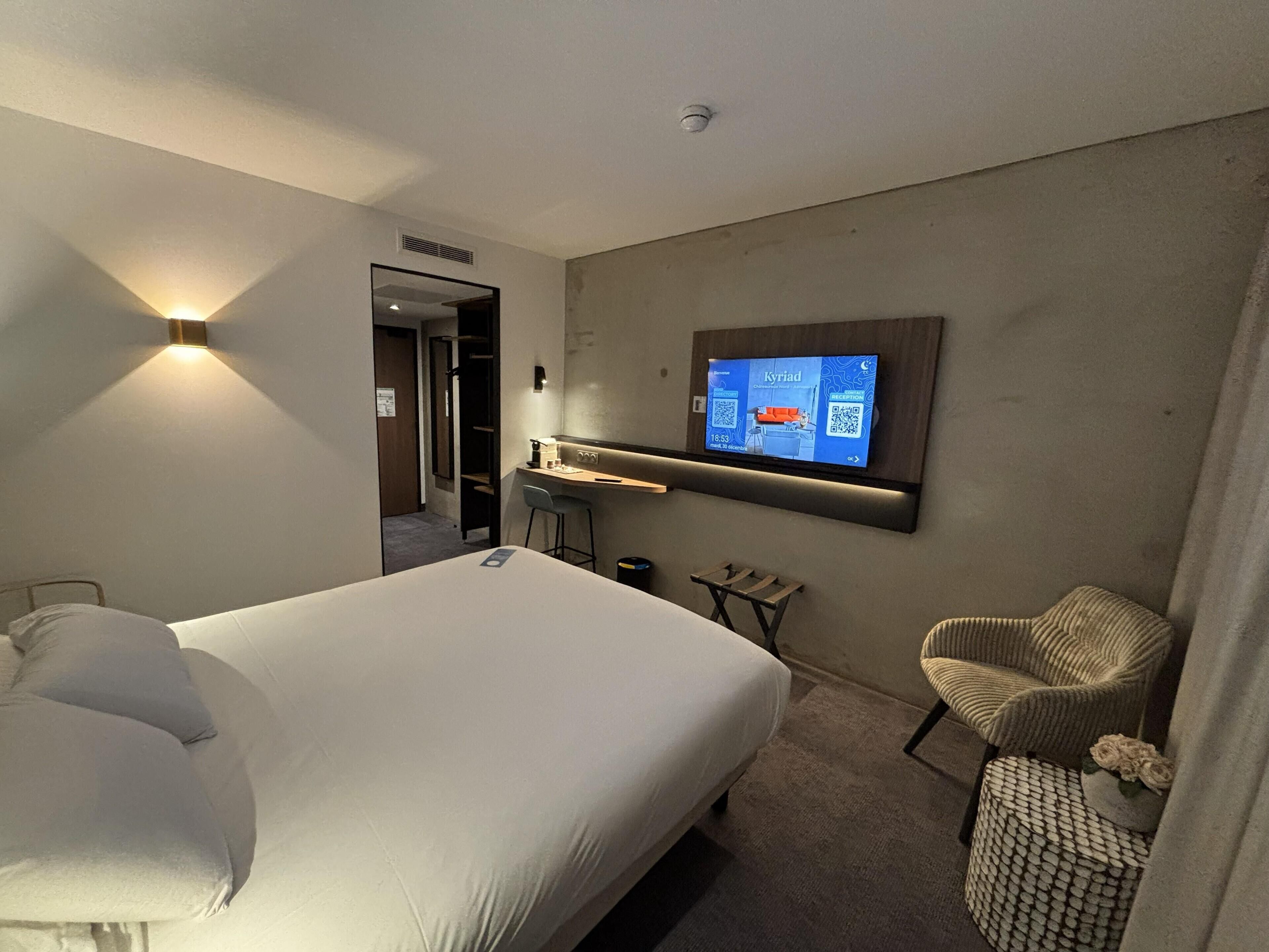 Ein Hotelzimmer mit Bett, Schreibtisch, Stuhl und an der Wand befestigtem Fernseher.
