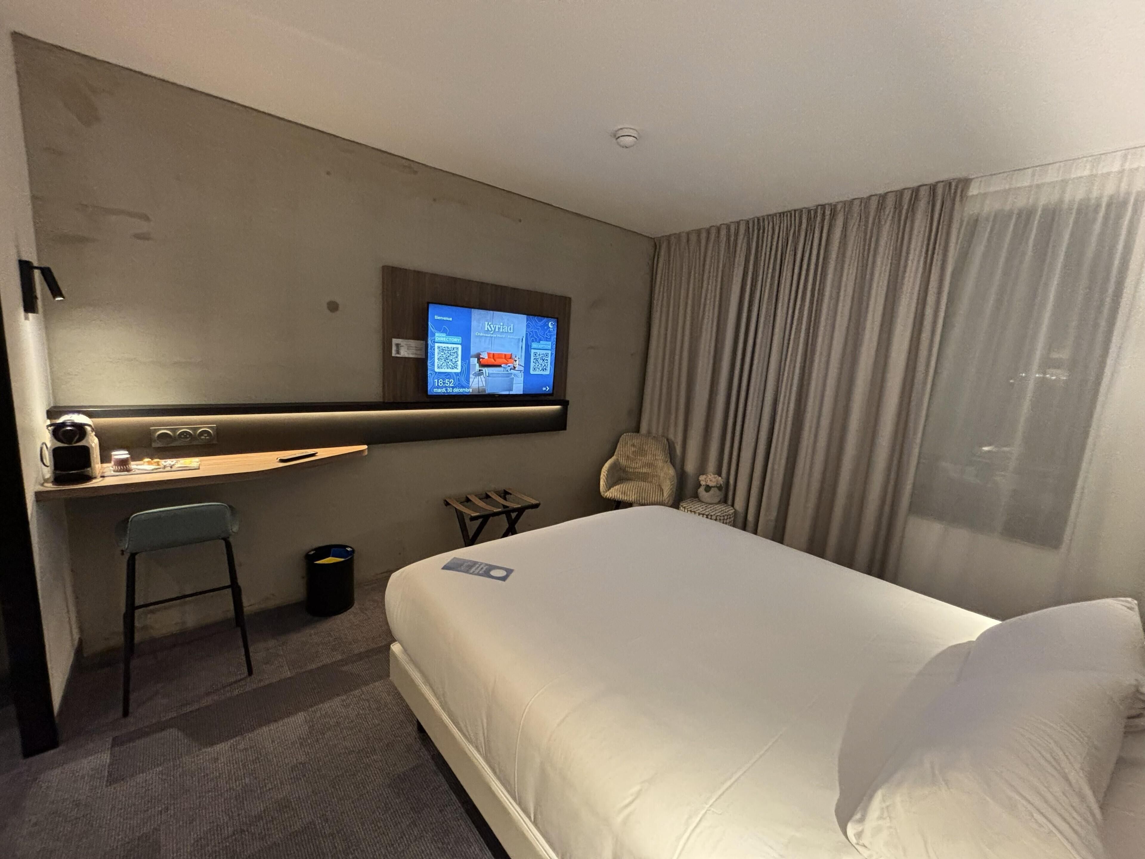 Una camera d'albergo con un letto, una scrivania con una sedia, un televisore fissato al muro e una finestra con tende.