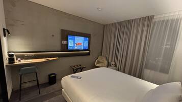 Una camera d'albergo con un letto, una scrivania con una sedia, un televisore fissato al muro e una finestra con tende.