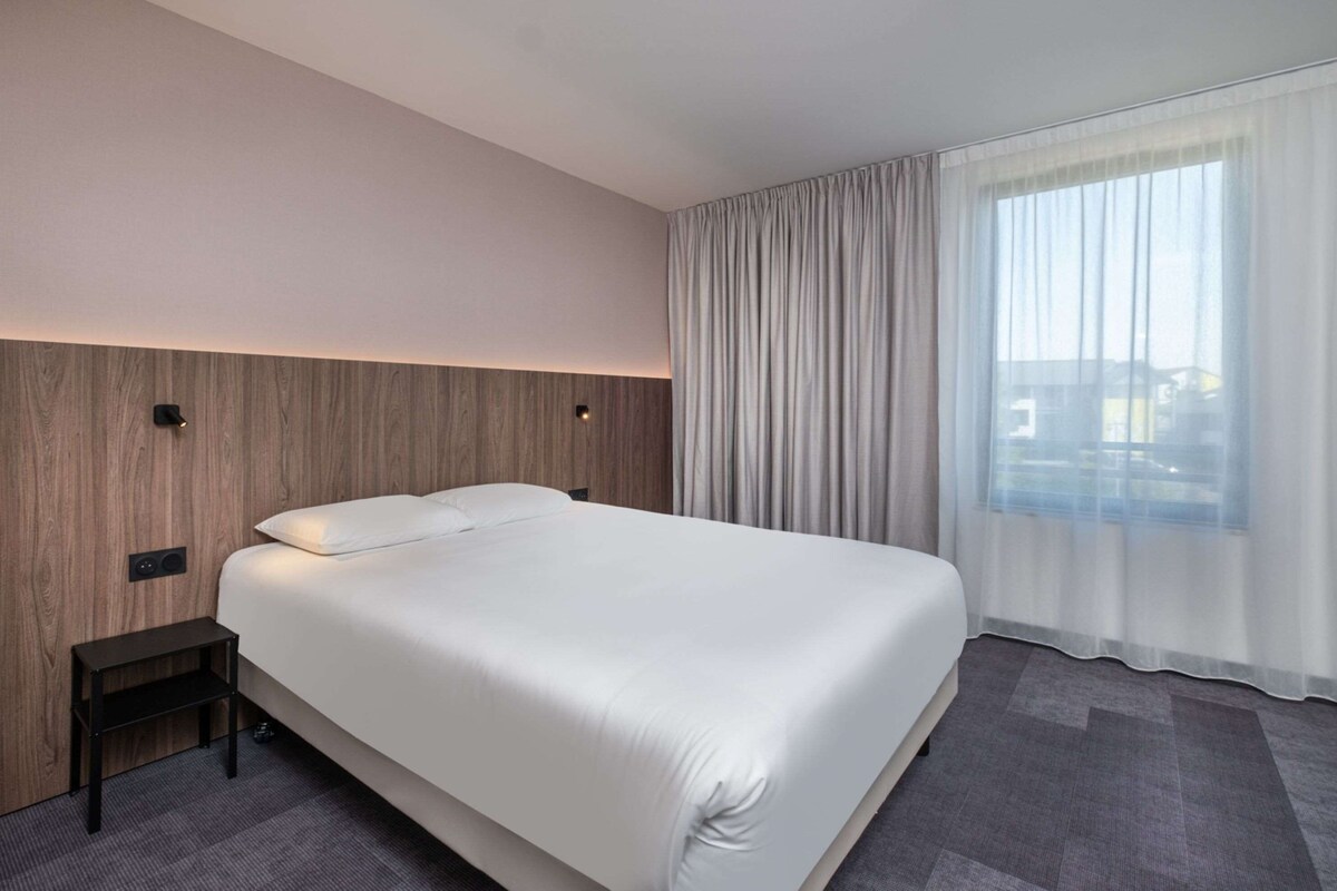 Une chambre d’hôtel moderne avec un grand lit, une table de chevet, un mur décoratif en bois et une grande fenêtre agrémentée de rideaux légers.