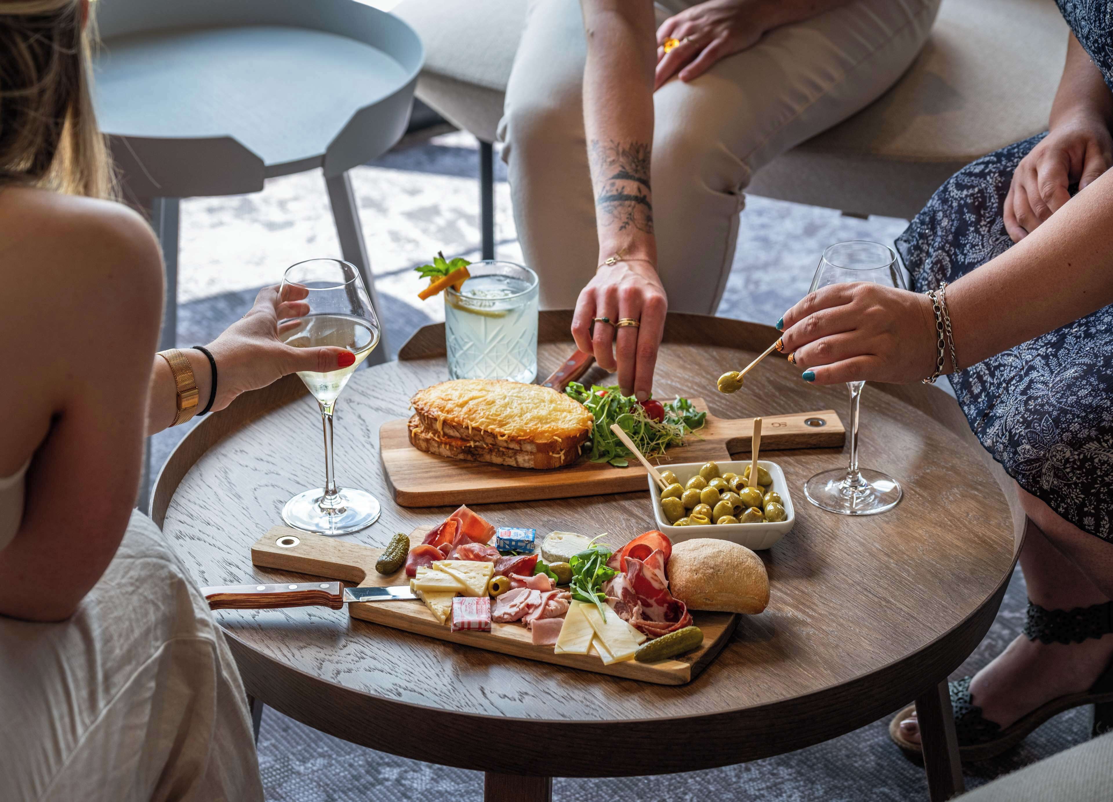 Une table ronde en bois, sur laquelle sont disposés divers aliments, notamment du fromage, des olives et de la charcuterie, ainsi que des verres à vin et un verre à eau.
