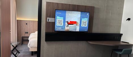 A hotel room with a bed, a desk, a chair, a TV mounted on the wall, and a door leading to another room.