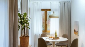 A dining area with a round table, four chairs, a bottle of wine, a basket with a plant, and a window with curtains.