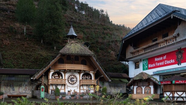 A traditional wooden building with a clock tower, surrounded by lush greenery and a mountainous backdrop.